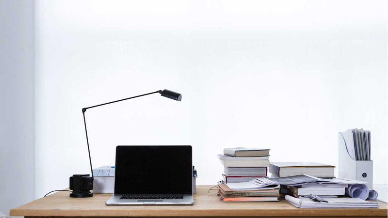 A typical modern office table with a laptop, lamp, books, and papers on a white wall background
